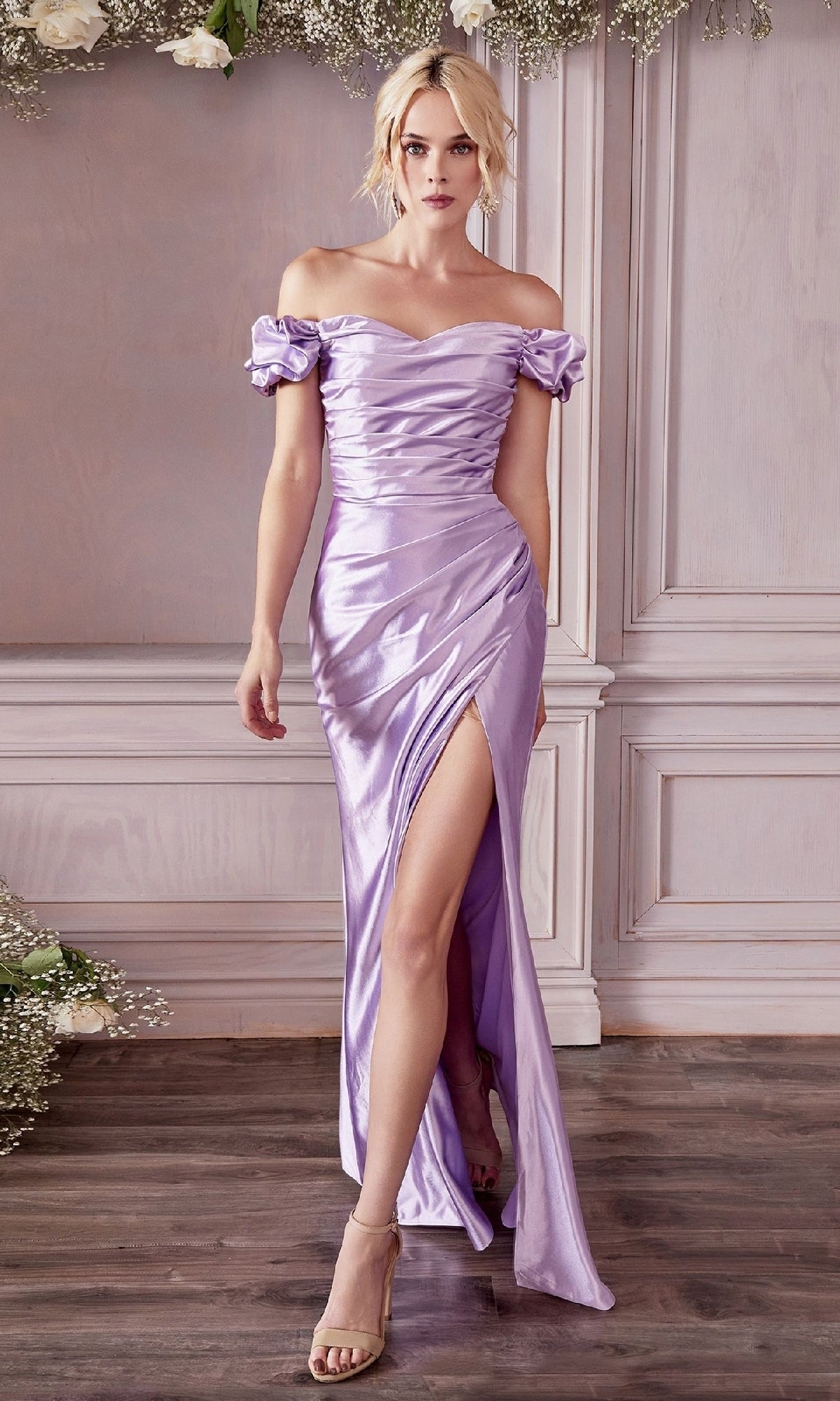 A woman stands indoors on a wooden floor, wearing the Ladivine KV1056 Ruched Long Satin Prom Dress in shiny lavender with off-the-shoulder puff sleeves and a high slit.